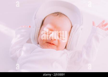 Das Kind trägt Kopfhörer. Frohes Baby. Ein neugeborenes Baby. Weiß auf Weiß Stockfoto