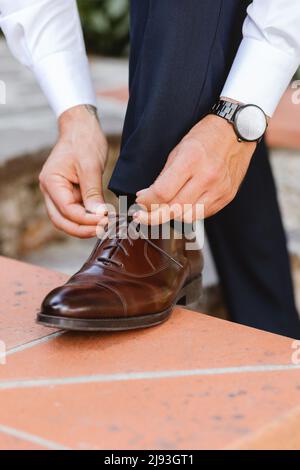 Ein Geschäftsmann oder Bräutigam ist in einem Außenbereich mit klassisch eleganten Schuhen verkleidet. Nahaufnahme der Hände und des Schuhs. Nahaufnahme der männlichen Hände, die schön schnürten Stockfoto