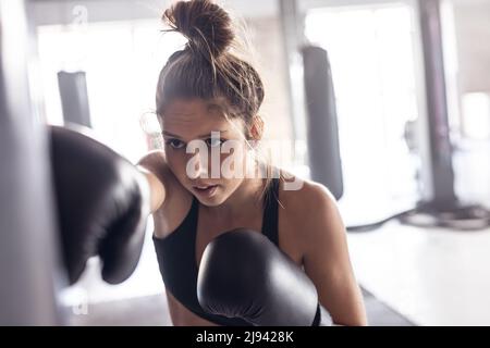 Selbstbewusste kaukasische junge Boxerin mit schwarzen Handschuhen, die im Fitnessstudio einen Boxsack trifft Stockfoto