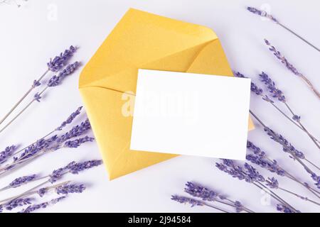 Ein weißes Einladungs- oder Grußkartenmockup mit Lavendel auf violettem Hintergrund und goldenem Umschlag. Kartenvorlage Stockfoto