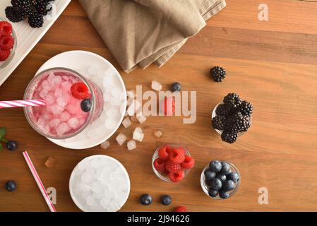 Fruchtgetränk mit Brombeeren, Himbeeren und Blaubeeren auf Holztisch mit Behältern voller Obst und Eis. Draufsicht. Horizontal komposit Stockfoto