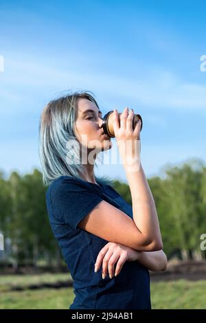 Blauhaarige Frau trinkt Kaffee zum Mitnehmen im Freien vor dem Hintergrund der Natur. Stockfoto