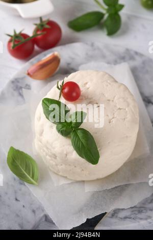 Ricotta-Käse. Hausgemachter Ricotta-Käse mit Basilikum, Knoblauch, Tomaten und grünen Oliven auf Pergamentpapier und Ständer. Italienisches Essen. Selektiver Fokus. Stockfoto