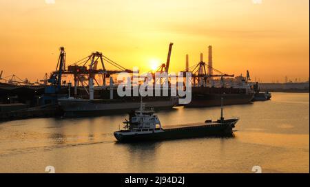 Schiffe im Hafen und auf der Durchreise transportieren und entladen verschiedene Fracht mit einem warmen schönen Sonnenaufgang im Hintergrund. Stockfoto