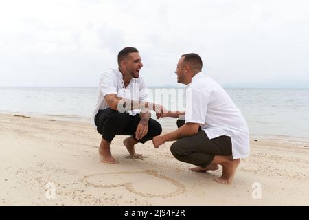 Schwule Paare halten die Hände neben einem Herz, das in den Sand an einem tropischen Strand gezogen wird Stockfoto