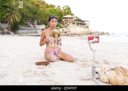 Streamer trinkt einen Kokosnusssaft, während sie am Strand mit einem Handy leben Stockfoto