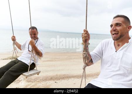 Porträt eines schwulen Paares, das auf einer Schaukel mitten am Strand schwingt Stockfoto