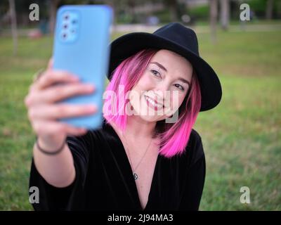 Frau mit schwarzem Hut und rosafarbenem Haar beim Selfie im Park Stockfoto