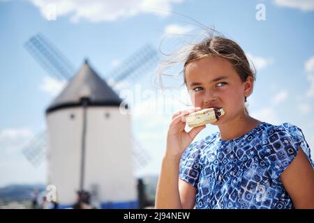 Niedliche kleine gebräunte 10s Mädchen in blauem Kleid essen Eis posiert im Freien, Consuegra Windmühlen auf dem Hintergrund. Sommer Süßigkeiten und Reisekonzept. Castil Stockfoto