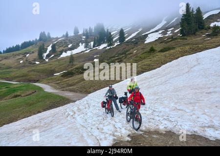 Eine Gruppe von drei aktiven Senioren, die mit ihren elektrischen Mountainbikes unterwegs sind und ein Schneefeld in den Allgäuer Alpen über Balderschwang, Bayern, Österreich, durchqueren Stockfoto