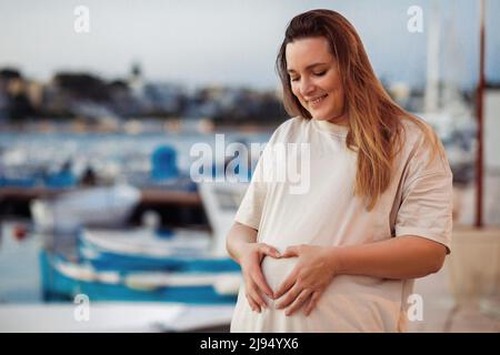 Fröhliche und ruhige blonde langhaarige Schwangere, die lächelt, den Bauch anschaut und den Bauch berührt. Hände wie Herz, Babypflege Stockfoto