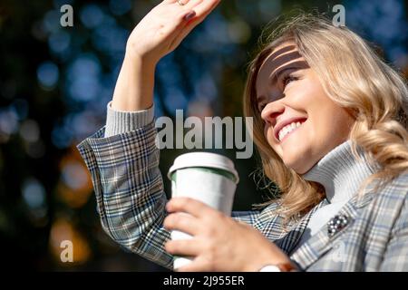 Lächelnd blonde in Tweed-Jacke mit einer Tasse Heißgetränk in den Händen, er blinzelt und schließt seine Hand vor direktem Sonnenlicht vor dem Hintergrund der Natur Stockfoto