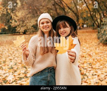 Zwei schöne Freundinnen in Strickpullover mit orangefarbenen Blättern in den Händen posieren im Herbstpark inmitten von herbstlichen Blättern. Frohe Feiertage. Gut gelaunt Stockfoto