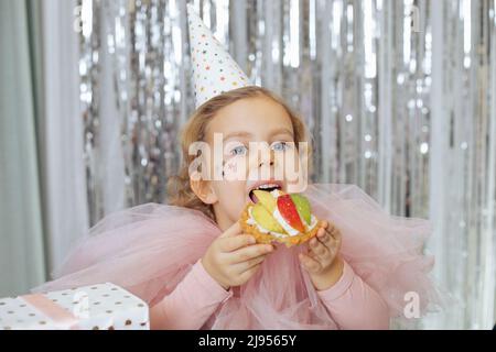 Das glückliche 4-5-jährige Mädchen im Partyhut im rosa Karnevalskleid wird Kuchen mit Schlagsahne und Obst probieren, die Geschenkbox steht neben ihr Stockfoto