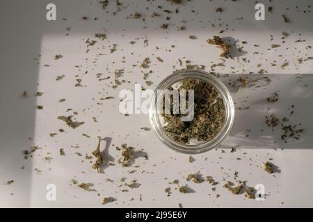 Unkrautladen. Glas mit getrockneten Cannabisknospen auf weißem Hintergrund mit zerkleinertem Unkraut. Stockfoto