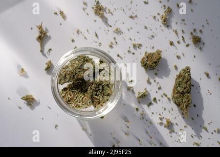 Unkrautladen. Glas mit getrockneten Cannabisknospen auf weißem Hintergrund mit zerdrücktem Marihuana. Stockfoto