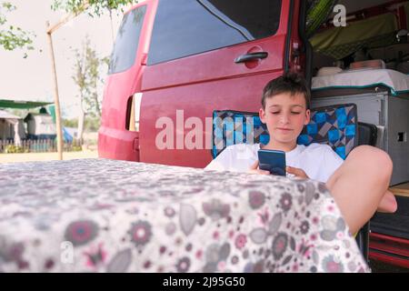 Junge mit Handy, der sich draußen vor dem Wohnmobil entspannen kann Stockfoto