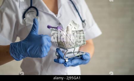 Ein gesichtsloser Arzt mit einem medizinischen Mantel und einem Stethoskop hält einen kleinen Einkaufswagen voller Medikamente. Apothekenkonzept. Der Apotheker zeigt einen Daumen Stockfoto