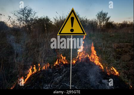 Brennendes trockenes Gras und Warnzeichen mit Ausrufezeichen warnen vor Gefahren im Feld mit Feuer. Konzept von Ökologie und Naturkatastrophen. Stockfoto