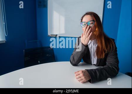 Eine Frau in Anzug und Brille macht im Konferenzraum Gähnen. Eine Büromanagerin mit chronischer Müdigkeit sitzt an einem Tisch in einer langweiligen Werkstatt Stockfoto