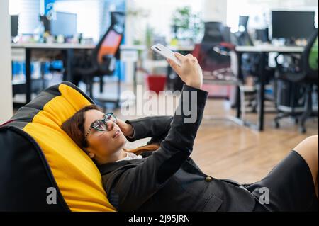 Eine Geschäftsfrau in einem Anzug ruht sich aus, während sie in einer bequemen Sitztasche sitzt und ein Selfie macht. Eine Mitarbeiterin nutzt ein Smartphone und entspannt sich Stockfoto