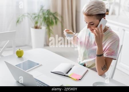 Trendige Frau mit Kaffeetasse, die auf dem Smartphone in der Nähe von leerem Notebook, Laptop und digitalem Tablet spricht Stockfoto