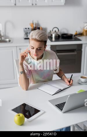 Stilvolle Frau, die in einem leeren Notebook schreibt und auf einem Mobiltelefon in der Nähe von Laptop und digitalem Tablet spricht Stockfoto