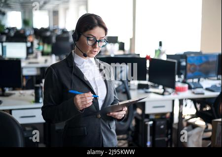 Eine seriöse Geschäftsfrau in einem Anzug beantwortet den Anruf des Kunden über das Headset. Die Managerin nimmt sich Notizen, während sie mit einem Kunden spricht. Administrator unter Stockfoto