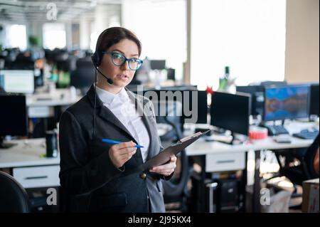 Eine seriöse Geschäftsfrau in einem Anzug beantwortet den Anruf des Kunden über das Headset. Die Managerin nimmt sich Notizen, während sie mit einem Kunden spricht. Administrator unter Stockfoto