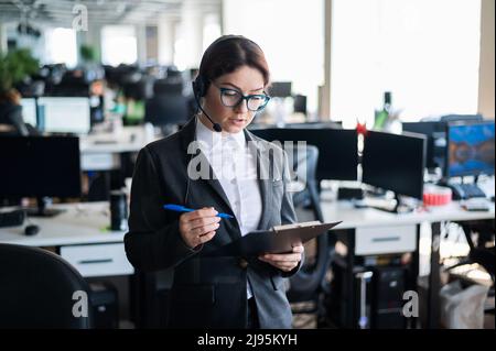 Eine seriöse Geschäftsfrau in einem Anzug beantwortet den Anruf des Kunden über das Headset. Die Managerin nimmt sich Notizen, während sie mit einem Kunden spricht. Administrator unter Stockfoto