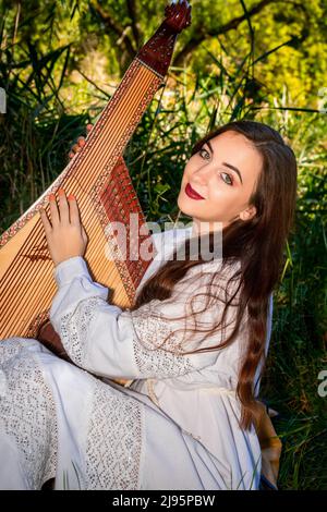 Die junge Ukrainerin in einem weißen Kleid und mit fließendem Haar sitzt auf dem grünen Gras und hält eine Bandura. Stockfoto