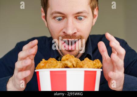 Hungriger, aufgerugter Mann, der mit gebratenen Hühnerflügeln auf einen Busket schaut. Stockfoto