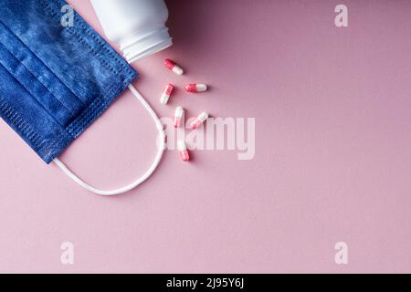 Mehrere Pillen und orale Kapseln auf einer medizinischen Einweg-Gesichtsmaske vor rosa Hintergrund. Stockfoto