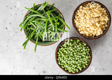 Tiefgefrorene grüne Bohnen, gefrorene Erbsen, gefrorener weißer Mais in einer Schüssel auf hellgrauem Hintergrund, flach liegend, Kopierraum Stockfoto