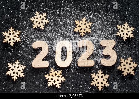 Konzept für das neue Jahr - Schneeflocken aus Holz und die Zahlen 2023, die mit Schnee auf schwarzem, glitzernden Sternenhintergrund bestreut wurden. Neujahrskarte. Stockfoto