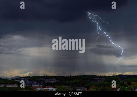 In der Ferne trifft ein Blitz auf einen Kommunikationsturm. Sehr dunkle Wolken mit starkem Regen von oben, Nachbarschaft unten ist ebenfalls dunkel. Platz für Text. Stockfoto