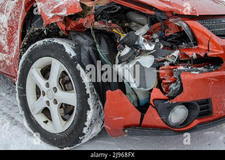 Vordere Ecke eines nicht identifizierten zertrümmerten Autos. Der größte Teil des Kotflügels fehlt. Gebrochener Kunststoff und Metall im Loch sichtbar. Schnee auf dem Boden und Reifen. Stockfoto