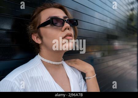 Nicht binärer Mann mit Sonnenbrille, der sich an das schwarze Tor auf der Straße lehnt, erinnert sich daran Stockfoto