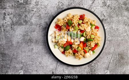 Nudelsalat mit Mozzarella, Grünpflanzen, Gurken und Tomaten auf einem runden Teller auf dunkelgrauem Hintergrund. Draufsicht, flach liegend Stockfoto