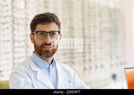 Gutaussehender lächelnder Augenarzt vor der Vitrine mit Brillen im optischen Laden Stockfoto