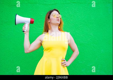 Eine rothaarige Frau mit Sonnenbrille und einem gelben Kleid hält ein Megaphon auf grünem Hintergrund. Stockfoto