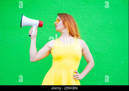 Eine rothaarige Frau mit Sonnenbrille und einem gelben Kleid hält ein Megaphon auf grünem Hintergrund. Stockfoto
