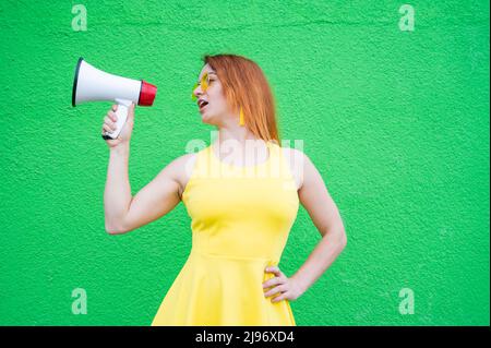 Eine rothaarige Frau mit Sonnenbrille und einem gelben Kleid hält ein Megaphon auf grünem Hintergrund. Stockfoto