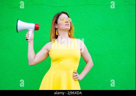Eine rothaarige Frau mit Sonnenbrille und einem gelben Kleid hält ein Megaphon auf grünem Hintergrund. Stockfoto