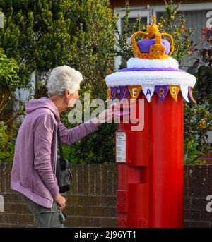 Weymouth, Dorset, Großbritannien. 21. Mai 2022. Ein Bewohner postet einen Brief an einer Briefbox in Weymouth in Dorset, die mit einer gehäkelten Staatskrone zur Feier des Platin-Jubiläums der Königin geschmückt wurde. Das Platin-Jubiläum von Elisabeth II. Wird vom 2. Juni bis 5. Juni 2022 im Vereinigten Königreich und im Commonwealth zum 70.. Jahrestag des Beitritts von Königin Elizabeth II. Am 6. Februar 1952 gefeiert. Bildnachweis: Graham Hunt/Alamy Live News Stockfoto