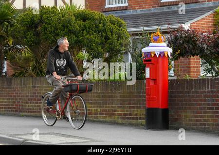 Weymouth, Dorset, Großbritannien. 21. Mai 2022. Ein Radfahrer passiert eine Briefbox in Weymouth in Dorset, die mit einer gehäkelten Staatskrone zur Feier des Platin-Jubiläums der Königin geschmückt wurde. Das Platin-Jubiläum von Elisabeth II. Wird vom 2. Juni bis 5. Juni 2022 im Vereinigten Königreich und im Commonwealth zum 70.. Jahrestag des Beitritts von Königin Elizabeth II. Am 6. Februar 1952 gefeiert. Bildnachweis: Graham Hunt/Alamy Live News Stockfoto