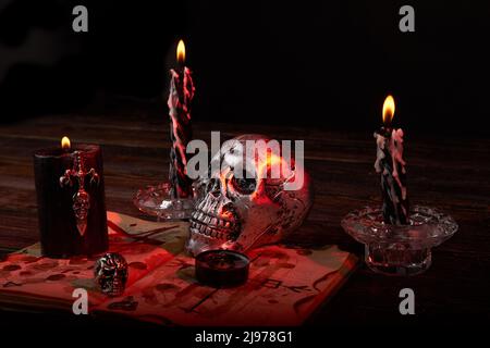 Stillleben Schädel mit Kerzen und andere Zubehör für sataische Ritual . Metallisches satanisches Kreuz und bibel verfärben sich mit Blut. Stockfoto