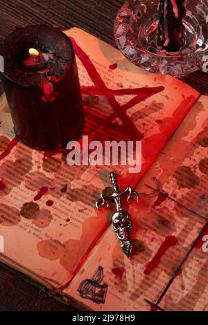 Vertikale Aufnahme des Antichristkreuzes auf der blutbefleckten satanischen bibel. Brennende schwarze Kerze auf dem Pentagramm. . Satanisches Wicca Ritualkonzept. Stockfoto