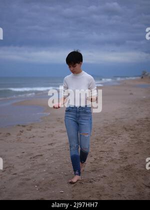 Transgender-Person, die barfuß auf dem Sand läuft, mit Muscheln in den Händen Stockfoto