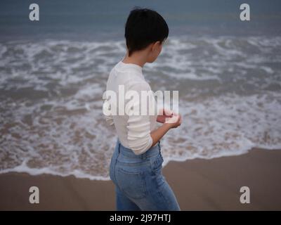 Frau, die vor dem Meer steht und die Wellen beobachtet Stockfoto
